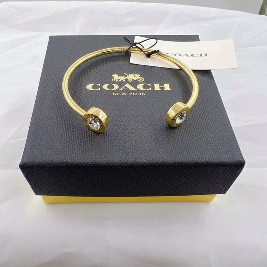 A gold-tone cuff bangle bracelet with clear crystal accents on both ends, displayed on top of a black Coach New York gift box with a white hangtag.
