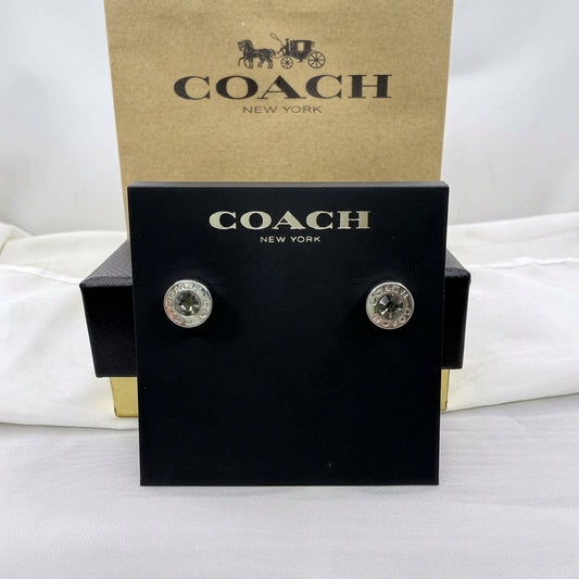 A pair of Coach stud earrings displayed on a black Coach New York card, with a brown Coach New York box behind it on a white fabric background.