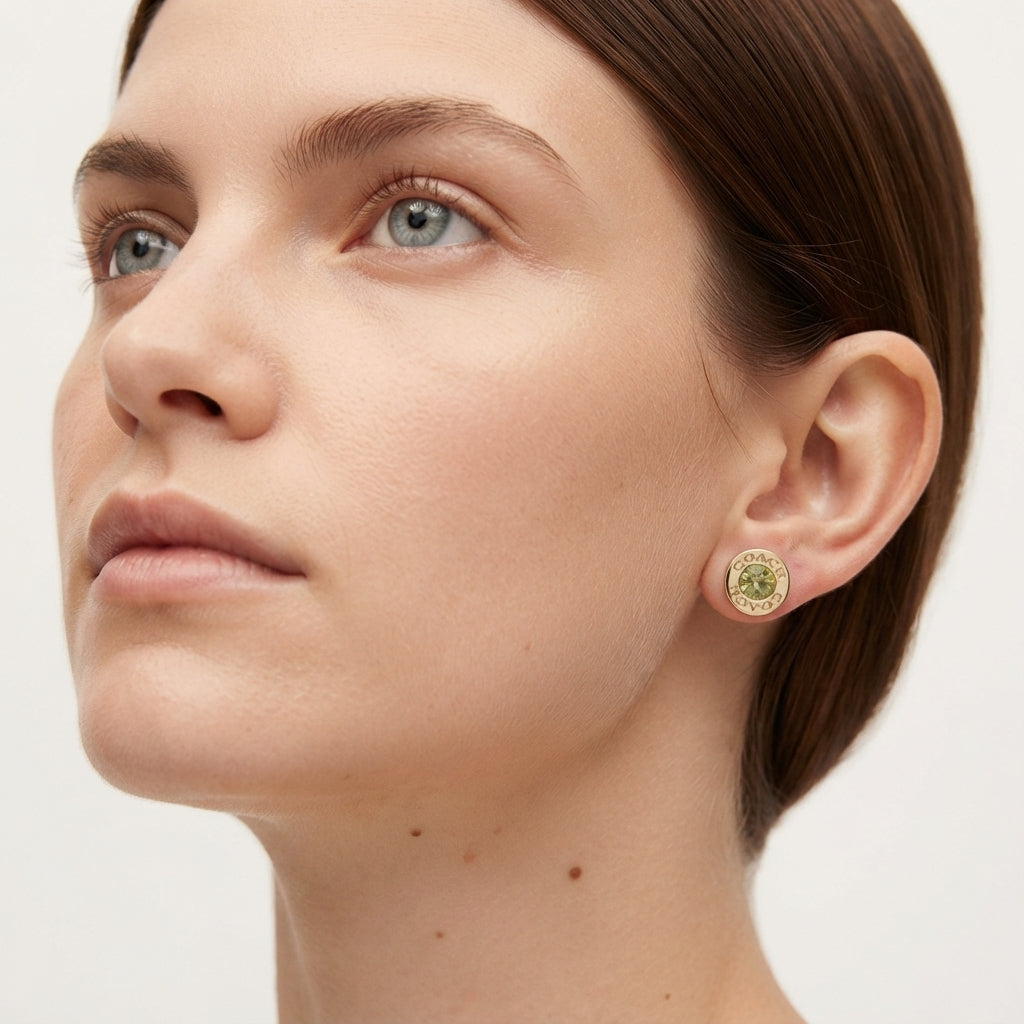 Close-up of a woman wearing a round gold-toned stud earring with a textured green center, shown against a clean light gray background.