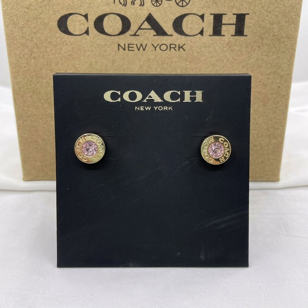 A pair of Coach stud earrings with gold-toned circular settings and pink gemstones, displayed on a black Coach New York card in front of a brown Coach box.