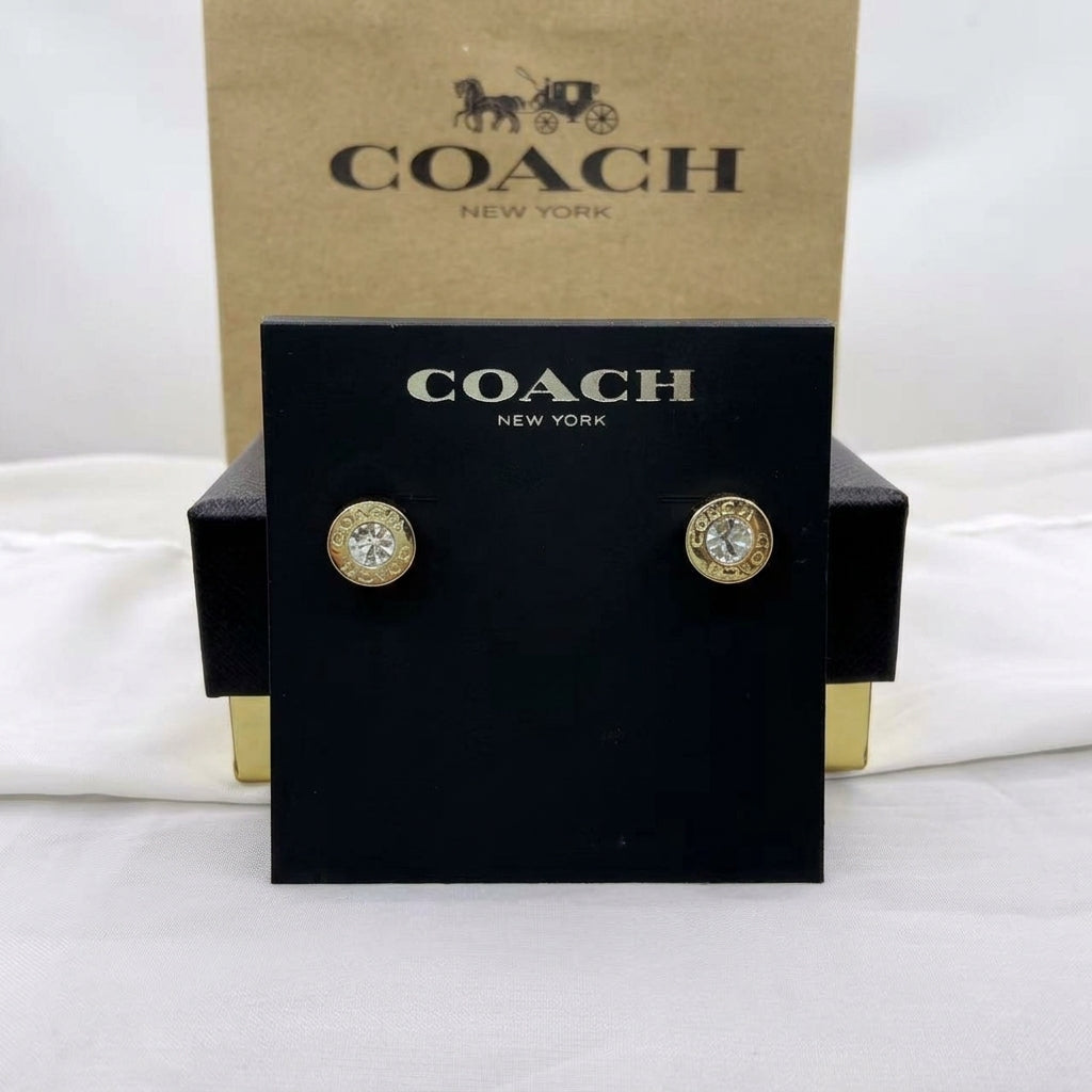 A pair of Coach New York stud earrings with gold-toned circular settings and clear stones, displayed on a black Coach New York card holder with a kraft Coach box behind it on a white fabric background.