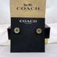 A pair of Coach New York stud earrings displayed on a black branded card, with the kraft Coach New York box behind it. The earrings have gold-tone circular settings with green faceted stones.