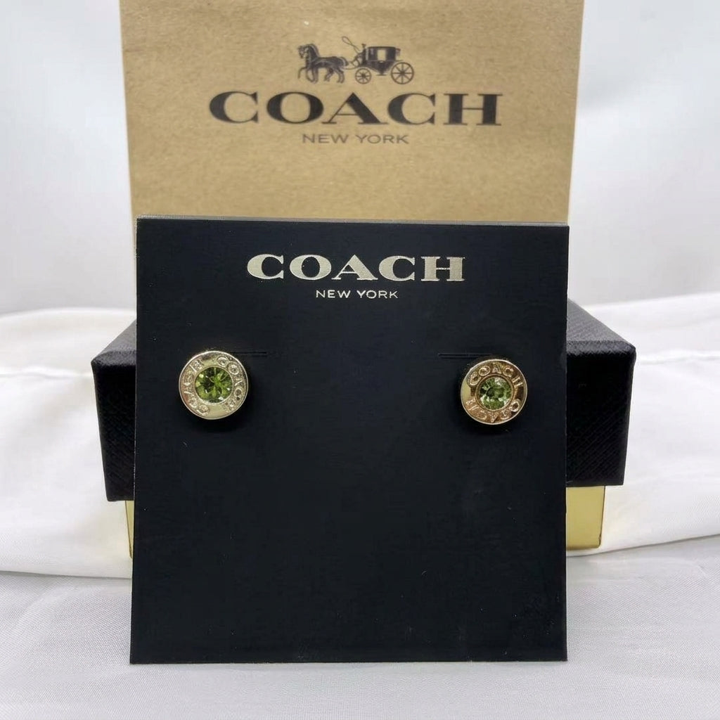 A pair of Coach New York stud earrings displayed on a black branded card, with the kraft Coach New York box behind it. The earrings have gold-tone circular settings with green faceted stones.