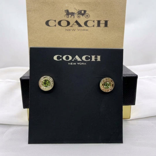 A pair of Coach New York stud earrings displayed on a black branded card, with the kraft Coach New York box behind it. The earrings have gold-tone circular settings with green faceted stones.