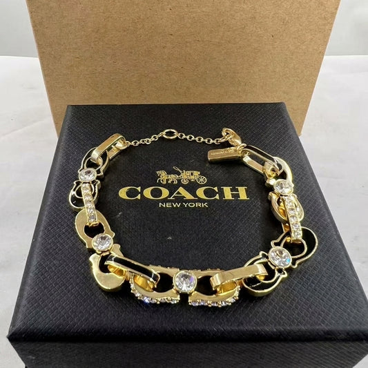 A gold-tone Coach New York bracelet with horsebit links and clear crystal accents, displayed on its black branded box.