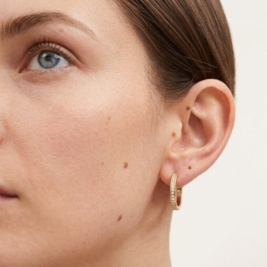 A close-up of a model's ear wearing a small gold hoop earring adorned with clear pavé stones, set against a clean white background.