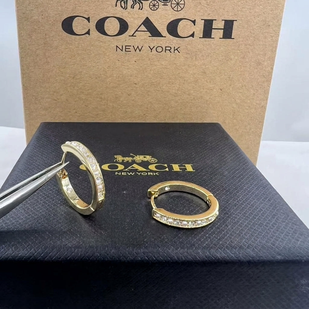 Gold hoop earrings with pavé-set clear stones, shown with tweezers next to a black Coach New York box and a brown Coach box in the background.