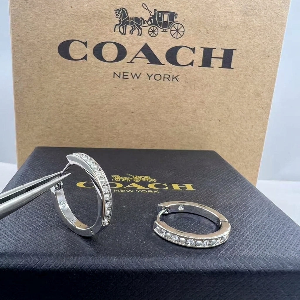 A pair of Coach huggie hoop earrings with clear pavé stones, shown with metal tweezers and the brand's signature brown box and black presentation box.