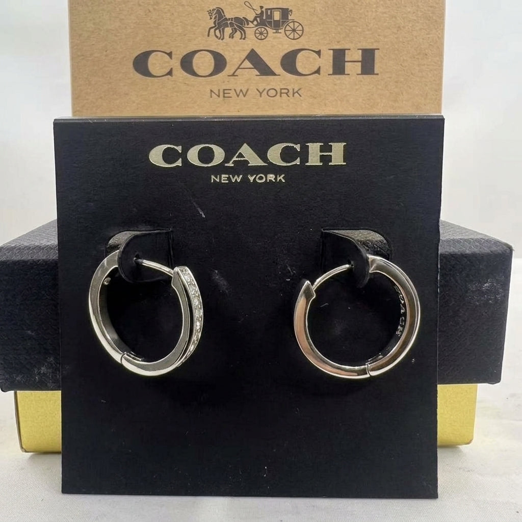 A pair of silver hoop earrings with small clear stones on one side, displayed on a black Coach New York card against a kraft Coach box.