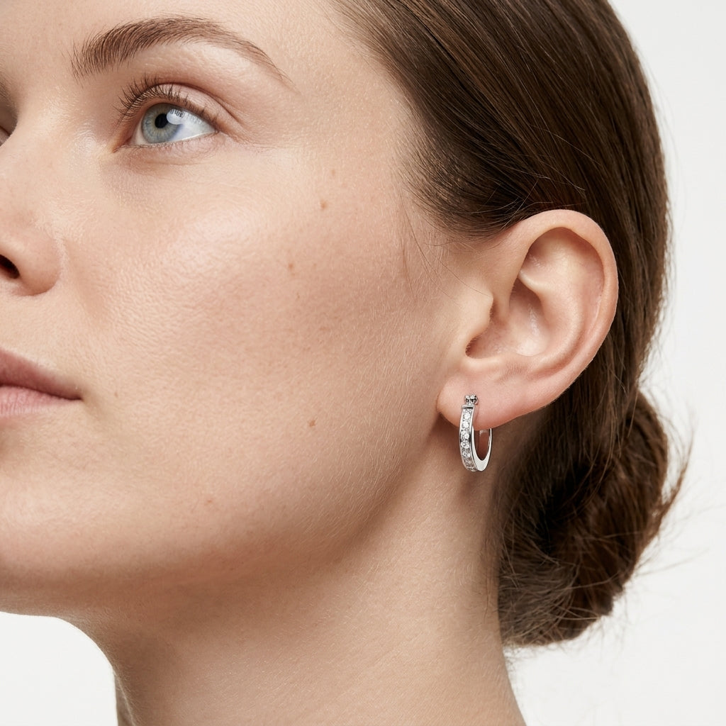 A close-up of a woman's ear wearing a small silver hoop earring set with clear stones, against a clean white background.