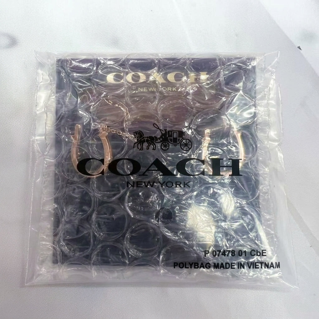 A Coach New York logo card inside a clear bubble wrap polybag, with gold-tone logo lettering and a horse-and-carriage emblem visible through the plastic.
