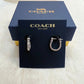 A pair of silver-tone Coach huggie hoop earrings displayed on a black branded card, with a dark blue Coach New York gift box behind it on a light stone surface.