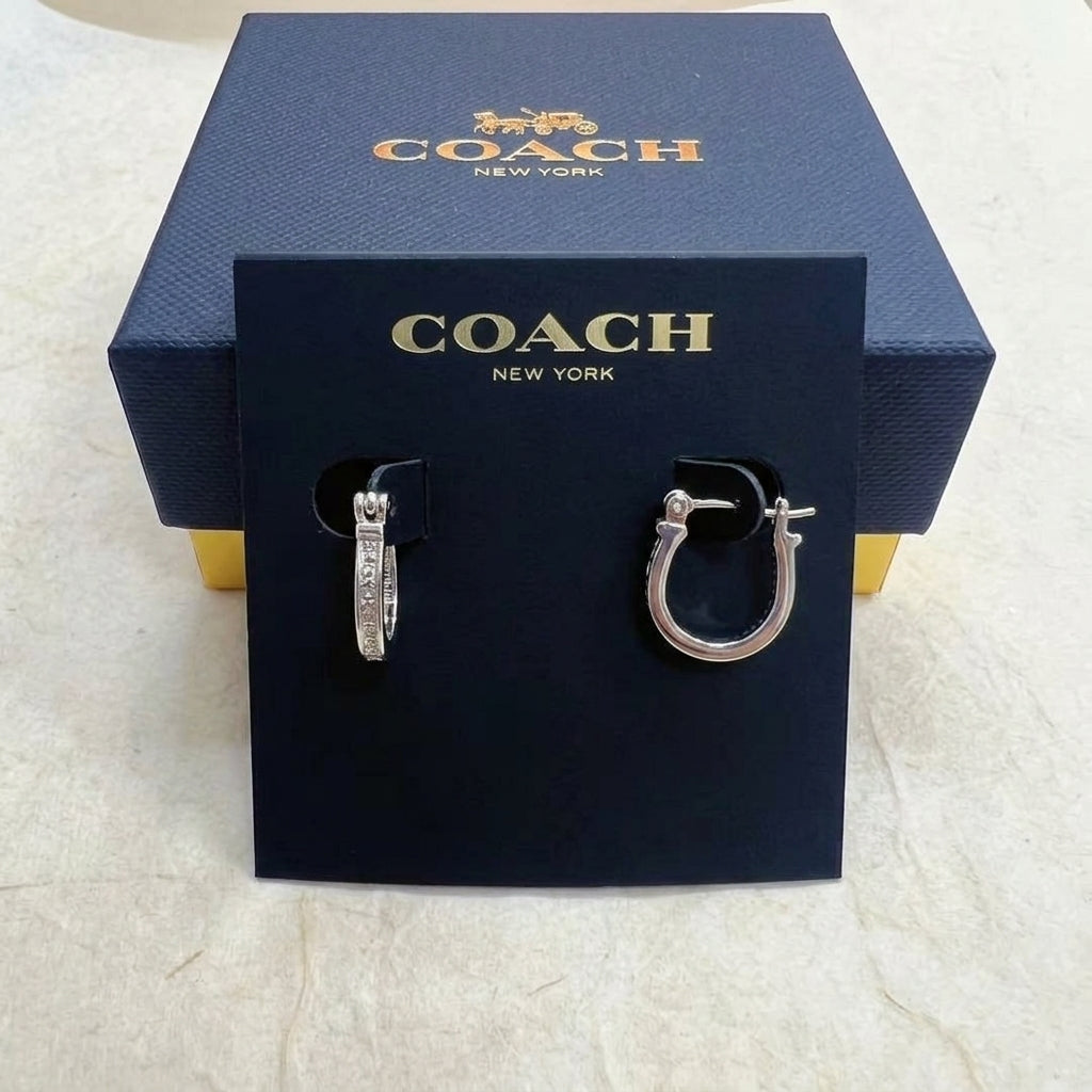 A pair of silver-tone Coach huggie hoop earrings displayed on a black branded card, with a dark blue Coach New York gift box behind it on a light stone surface.