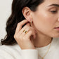A close-up lifestyle shot of a woman wearing a small gold hoop earring and a wide gold ring, with her hand near her ear. She is wearing a white ribbed sweater against a clean, light off-white background.