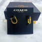 A pair of gold-tone hoop earrings displayed on a black Coach New York card, with the black Coach box behind it on a white fabric background.
