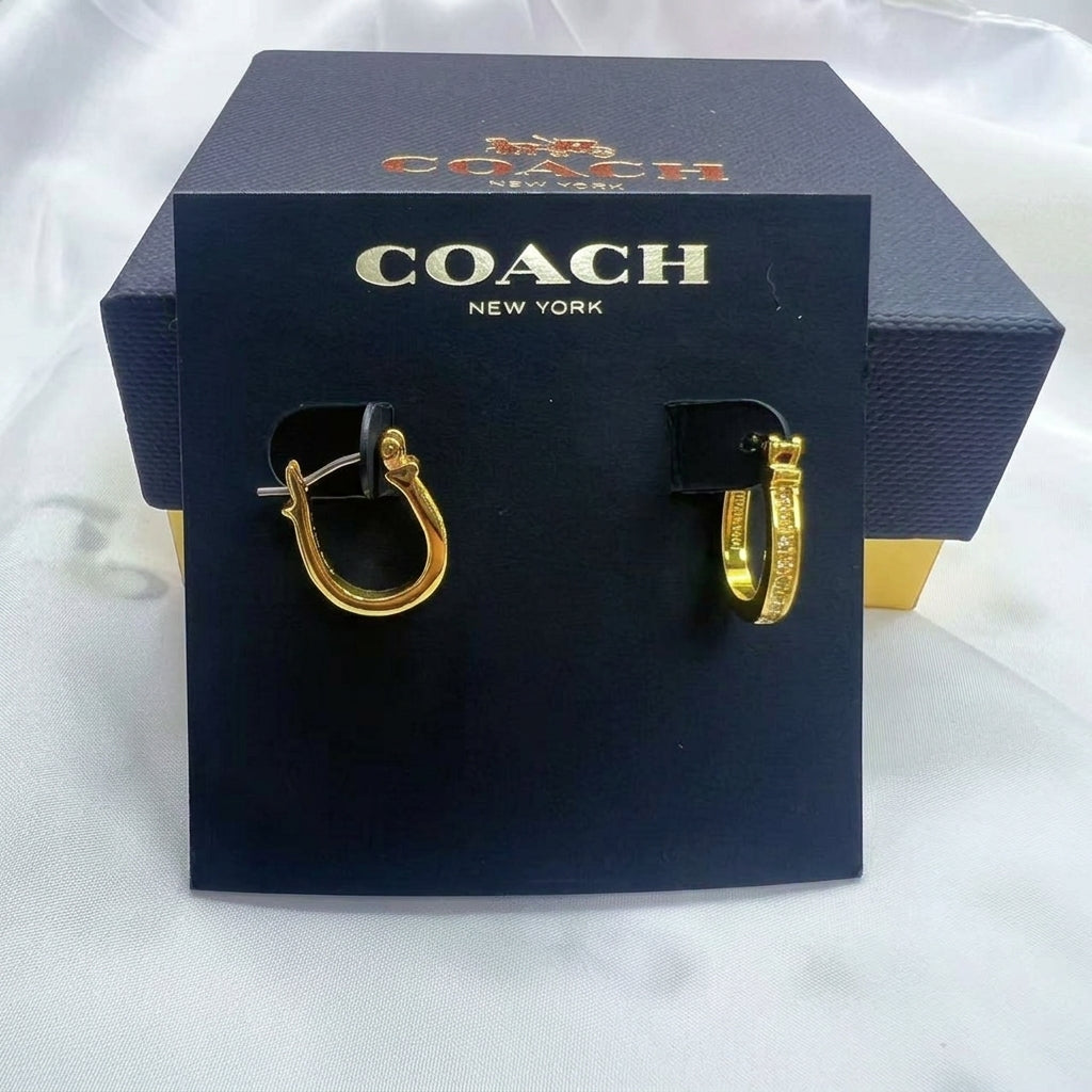 A pair of gold-tone hoop earrings displayed on a black Coach New York card, with the black Coach box behind it on a white fabric background.