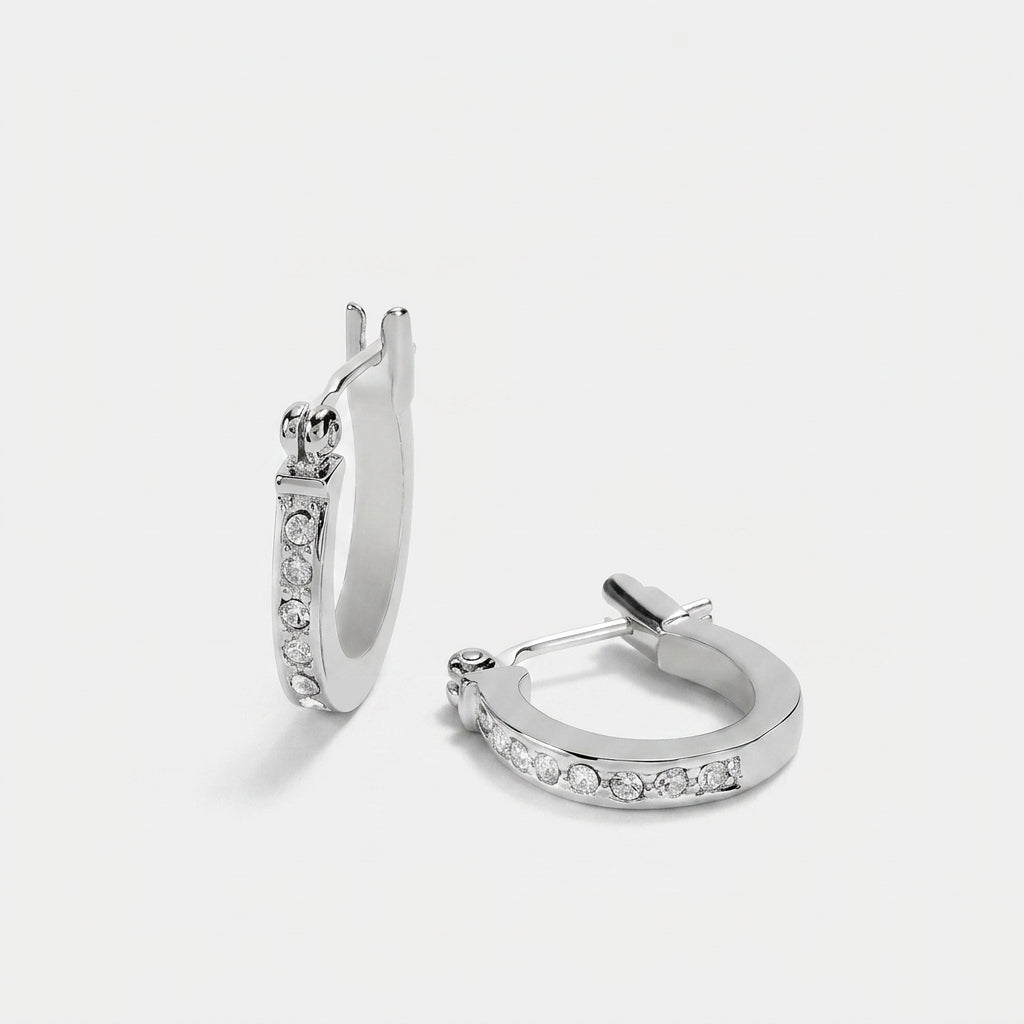 A pair of silver huggie hoop earrings with clear pavé-set stones, shown from two angles on a light gray background.