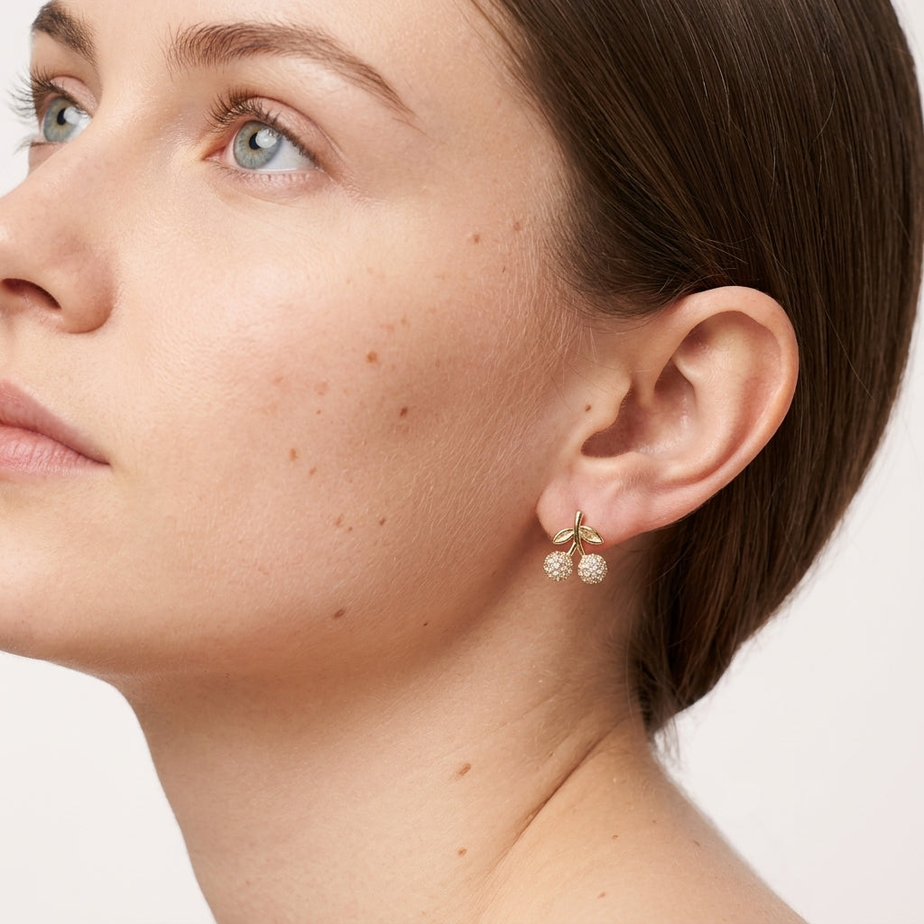 A close-up of a woman's ear wearing a small gold earring with two dangling pearl-like bead clusters, shown against a clean light background.