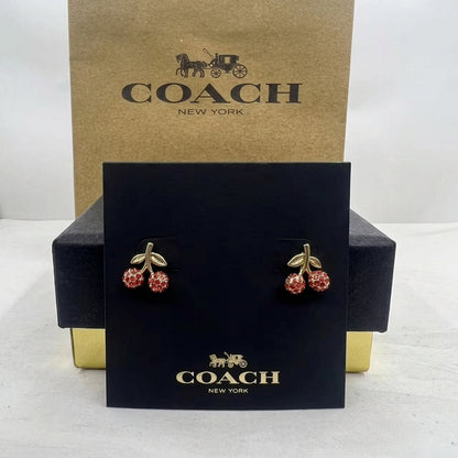 A pair of Coach New York cherry stud earrings with red crystal-like accents and gold-tone leaves, displayed on a black branded card in front of a Coach New York box.