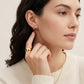 A close-up of a woman wearing a small gold and red berry earring, with a matching gold ring on her finger and a delicate gold necklace. The background is a soft, warm off-white.