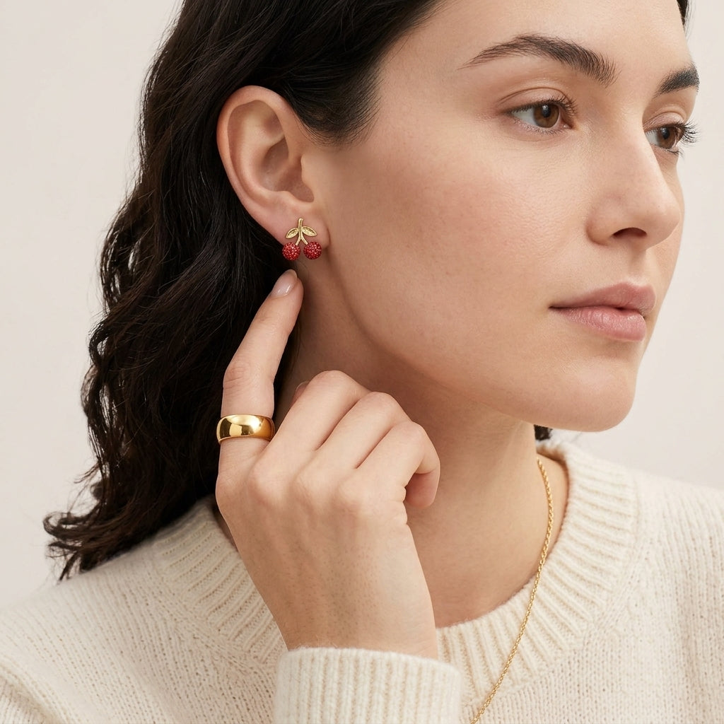 A close-up of a woman wearing a small gold and red berry earring, with a matching gold ring on her finger and a delicate gold necklace. The background is a soft, warm off-white.