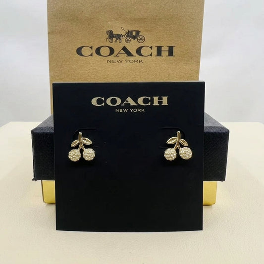 A pair of Coach New York cherry stud earrings with gold-toned stems and clear crystal-like round stones, displayed on a black Coach card with a brown Coach box behind it.