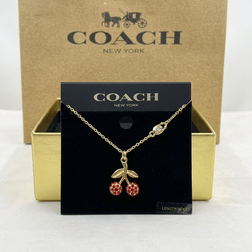 A gold-tone Coach necklace with a cherry pendant is displayed on a black Coach New York card, with the gold Coach box behind it on a white surface.