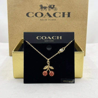 A gold-tone Coach necklace with a cherry pendant is displayed on a black Coach New York card, with the gold Coach box behind it on a white surface.