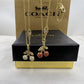 Two gold-tone Coach necklaces displayed on a black presentation box in front of a brown Coach box. One necklace has clear crystal-embellished cherries, and the other has red crystal-embellished cherries, both with gold leaves and chains.