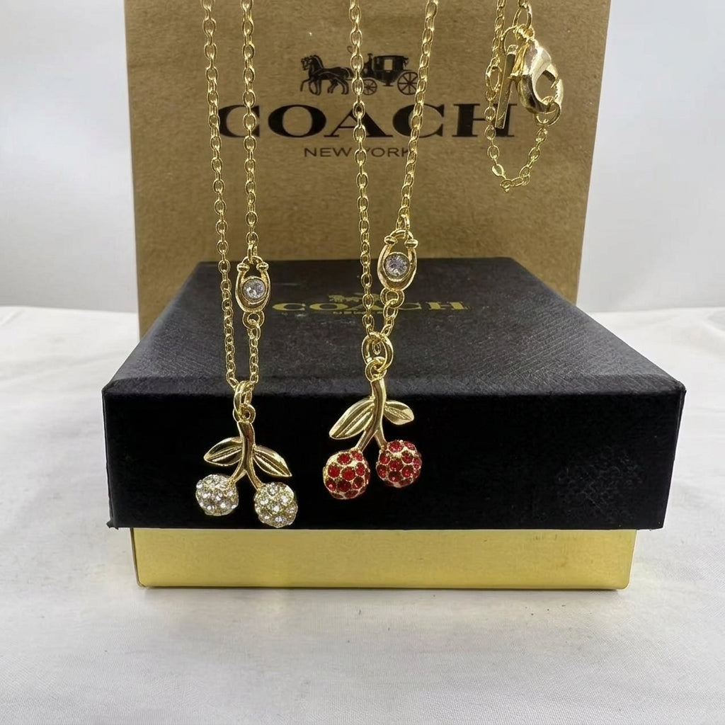 Two gold-tone Coach necklaces displayed on a black presentation box in front of a brown Coach box. One necklace has clear crystal-embellished cherries, and the other has red crystal-embellished cherries, both with gold leaves and chains.