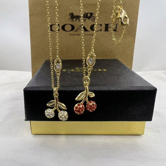 Two gold-tone Coach necklaces displayed on a black presentation box in front of a brown Coach box. One necklace has clear crystal-embellished cherries, and the other has red crystal-embellished cherries, both with gold leaves and chains.