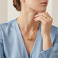 A close-up lifestyle shot of a model wearing a delicate gold chain necklace with a unique cylindrical pendant featuring textured gold and silver bands, paired with a matching gold ring on her finger.