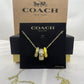 A Coach New York necklace displayed on a black branded card, with the gold-tone chain draped below. The pendant features stacked cylindrical charms in yellow, silver, and white with Coach logos.