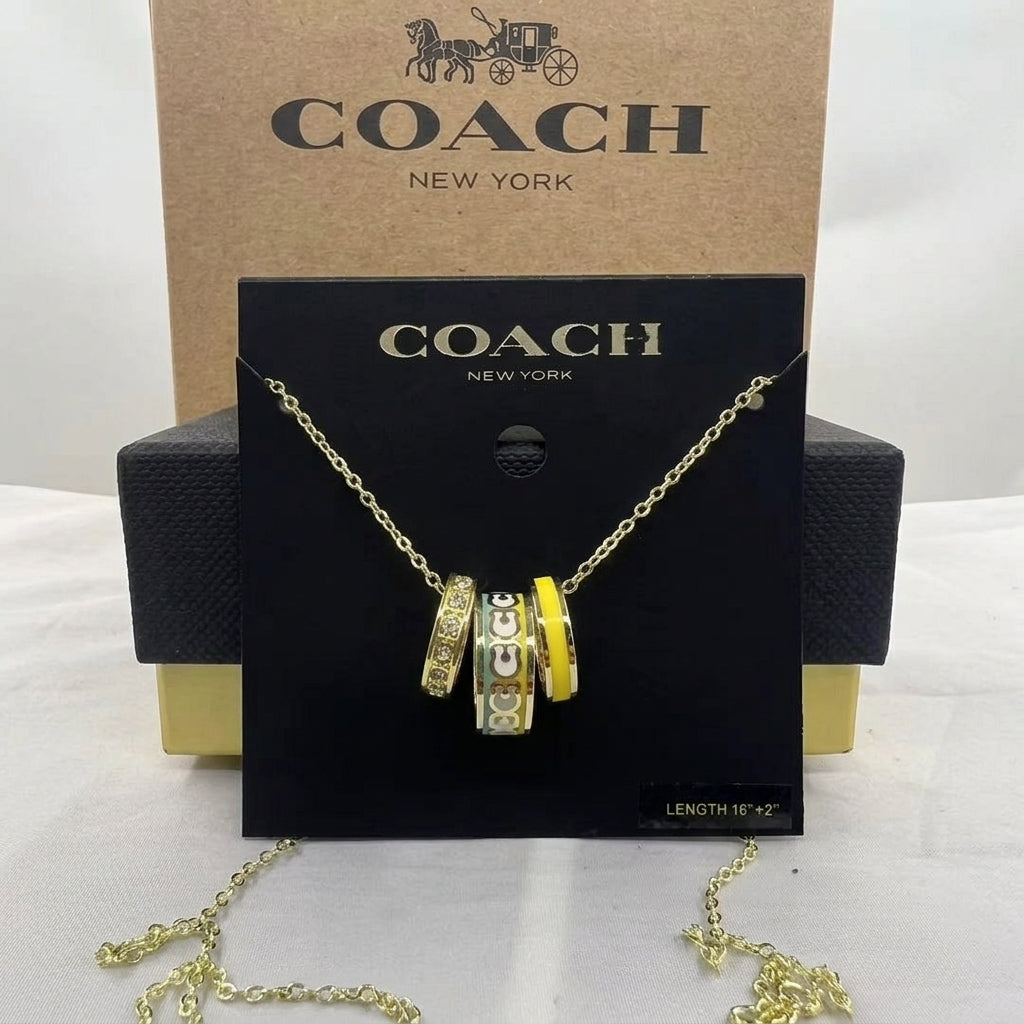A Coach New York necklace displayed on a black branded card, with the gold-tone chain draped below. The pendant features stacked cylindrical charms in yellow, silver, and white with Coach logos.
