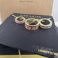 Three Coach New York chain rings displayed on a black branded box, with a kraft paper box behind them. The rings are gold-tone with colorful enamel-style bands in pink, orange, and red, and some have rhinestone detailing.