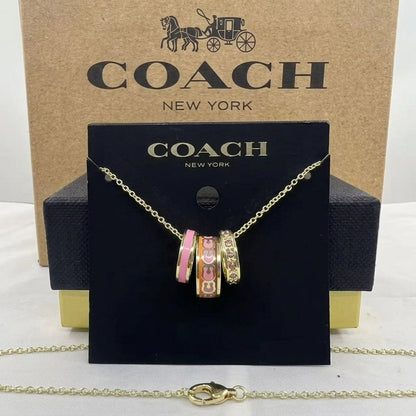 A Coach New York necklace displayed on a black branded card, with the gold-tone chain draped in front. The pendant features three charm-style rings with pink, peach, and gold details.
