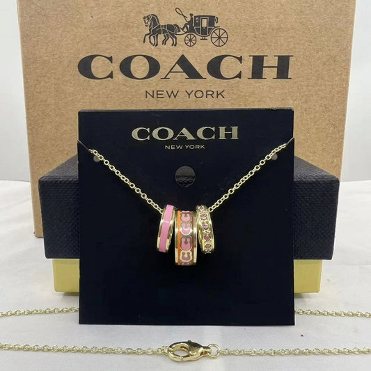 A Coach New York necklace displayed on a black branded card, with the gold-tone chain draped in front. The pendant features three charm-style rings with pink, peach, and gold details.