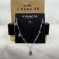 A Coach New York silver charm necklace displayed on a black branded card, with a kraft Coach box behind it on a white fabric surface.