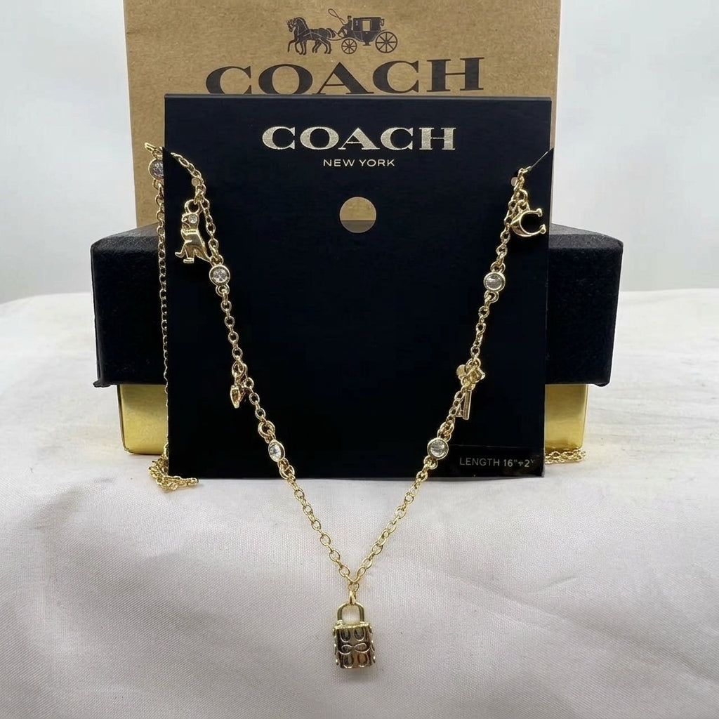 A gold-tone Coach necklace with a padlock pendant and small charm details, displayed on a black Coach New York card with a kraft Coach box behind it.