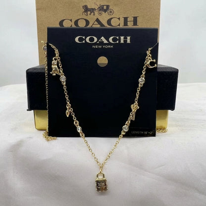 A gold-tone Coach necklace with a padlock pendant and small charm details, displayed on a black Coach New York card with a kraft Coach box behind it.