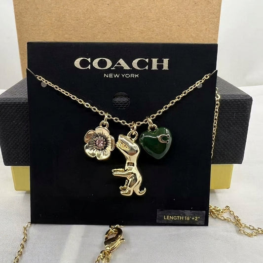 A Coach New York necklace displayed on a black branded card, featuring a gold-tone chain with a flower charm, a dinosaur charm, and a green heart charm. The necklace is shown in front of a black and gold Coach box.