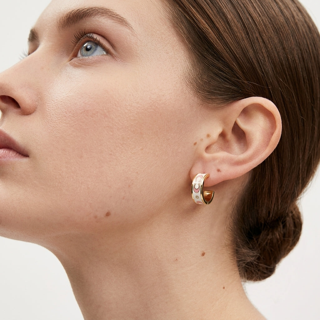 A close-up of a woman's ear and side profile wearing a small gold huggie hoop earring with a white enamel inlay and a tiny pink gemstone.