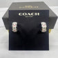 A pair of Coach New York hoop earrings displayed on a black branded card, with the black Coach box behind it on a white fabric surface.
