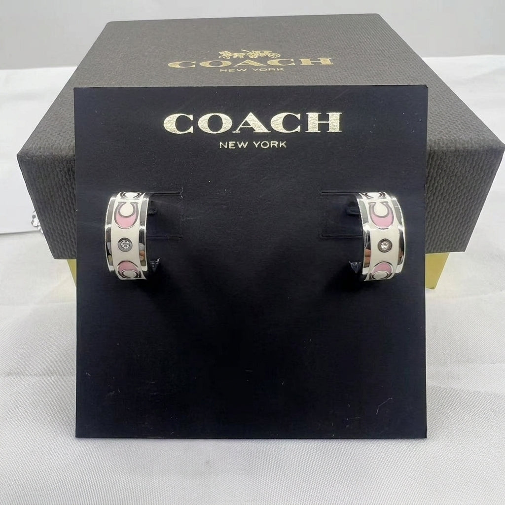 A pair of Coach New York hoop earrings displayed on a black branded card, with the black Coach box behind it on a white fabric surface.