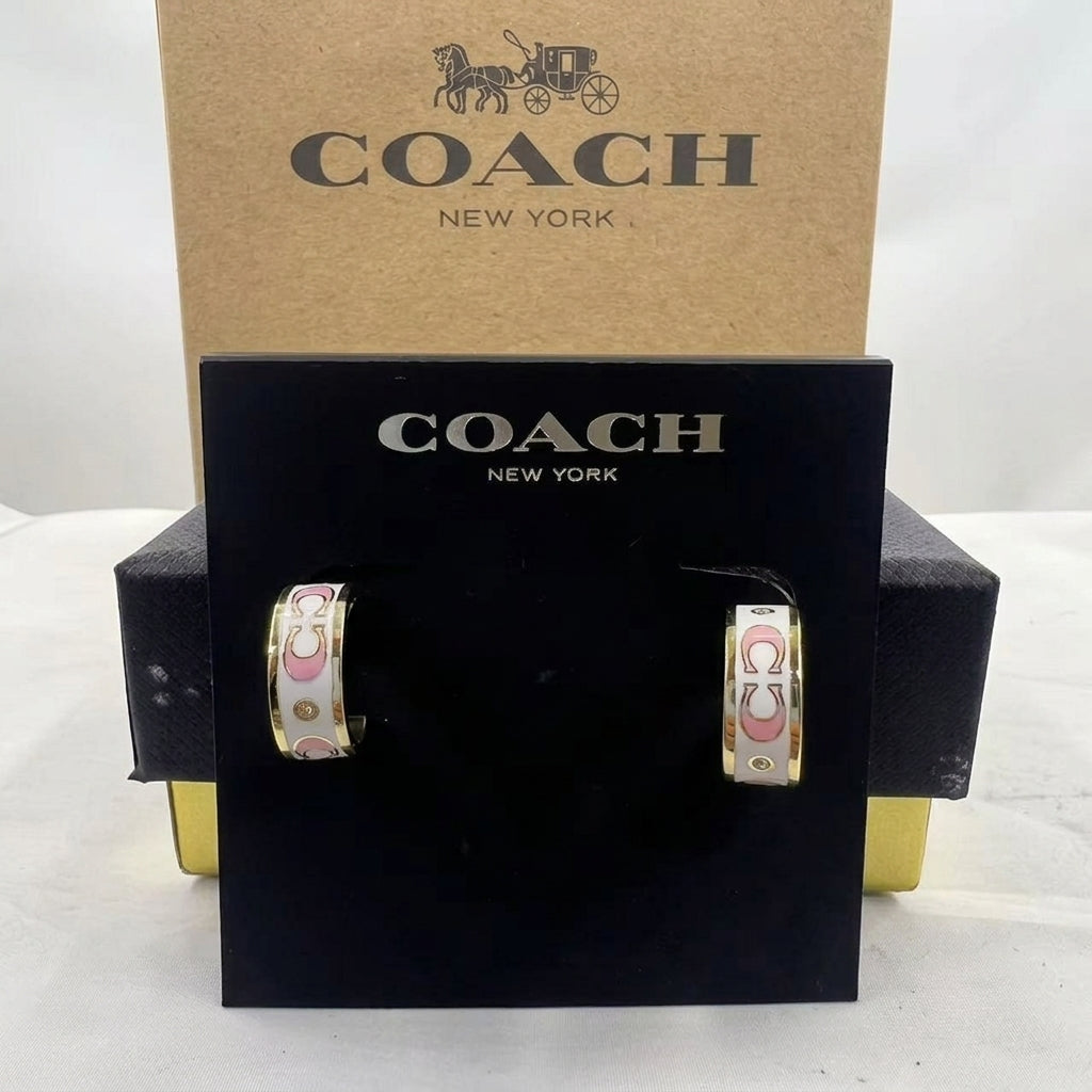 A pair of Coach New York hoop earrings displayed on a black branded card, with the kraft Coach box behind them on a white studio background.