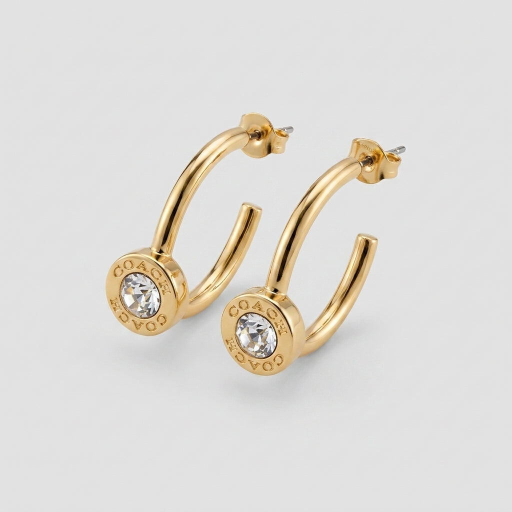 A pair of gold hoop earrings with a circular disc at the end featuring a clear crystal and the brand name 'COACH' engraved on it, set against a light gray background.