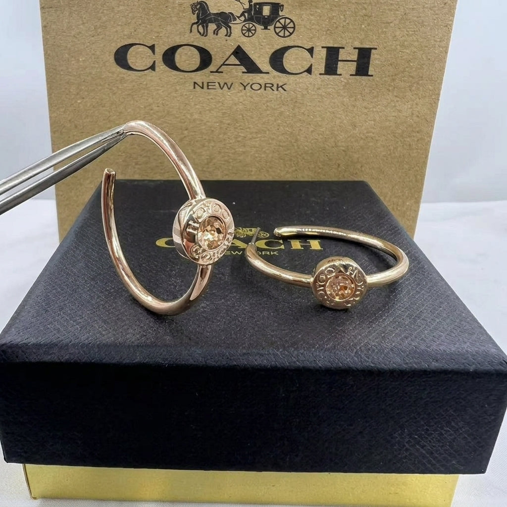 Two Coach New York gold-tone bangle bracelets with circular floral medallions, displayed on a black box with the Coach New York box behind them. One bracelet is held by tweezers.