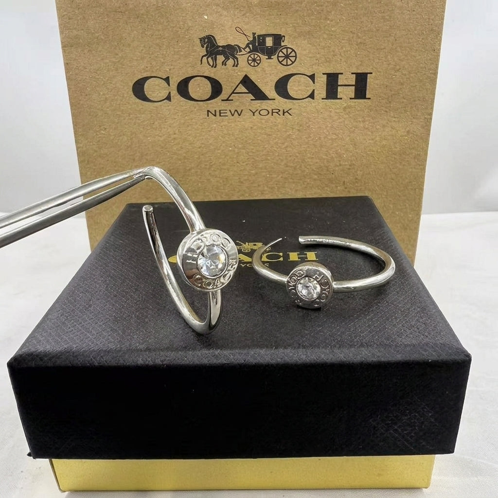 A pair of silver-tone Coach New York ring bangles with round crystal-encrusted bezels, displayed on a black box with a gold base in front of a brown Coach New York box.
