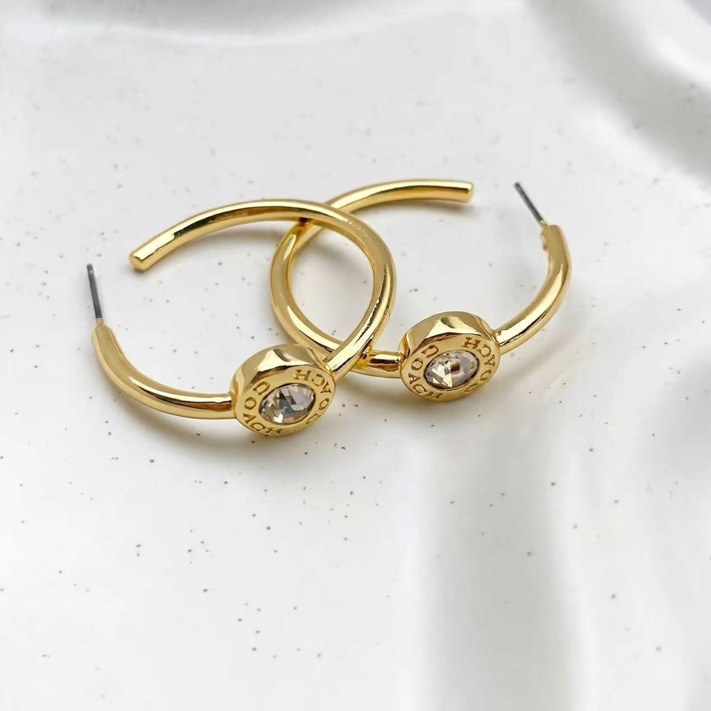 A pair of gold hoop earrings with circular disc charms featuring clear rhinestones and the word 'HOKYO' engraved on the side, displayed on a white textured surface.
