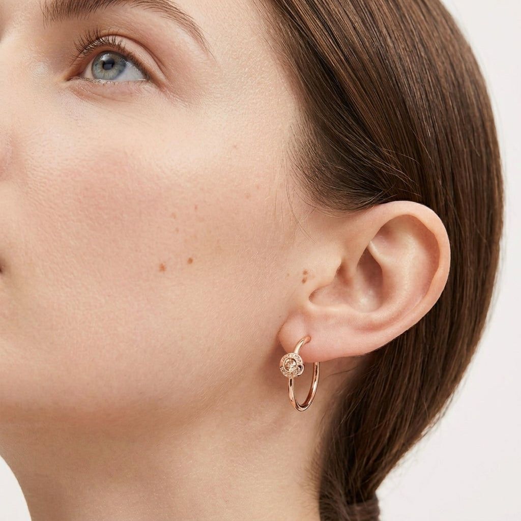 A close-up of a woman's ear wearing a delicate rose gold huggie hoop earring with a small clear gemstone at the front, set against a clean white background.